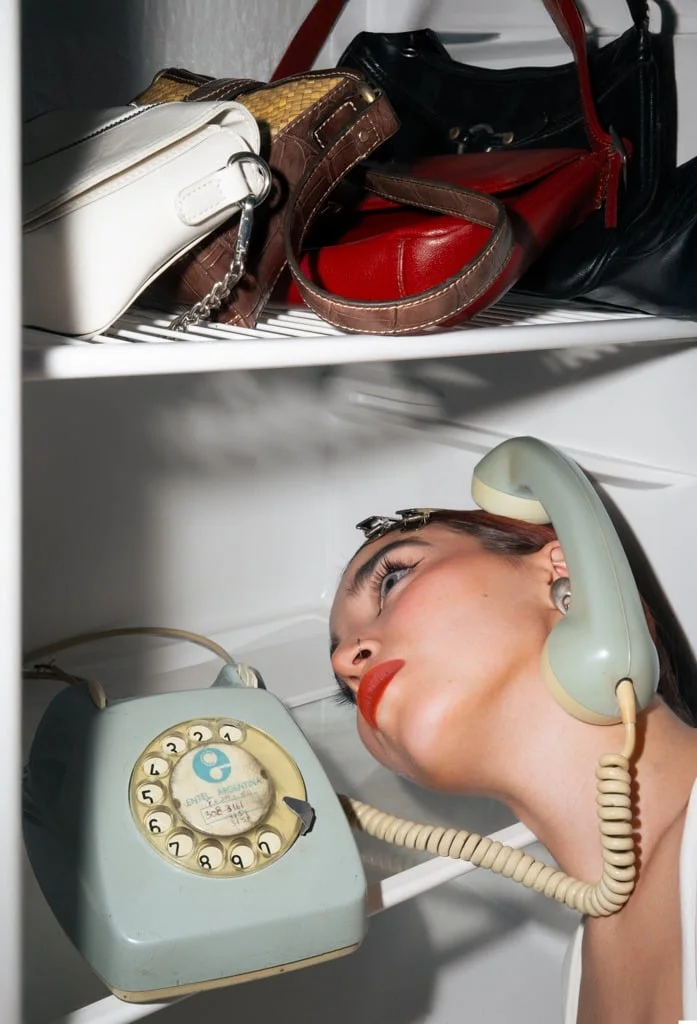 Modelo recostada dentro de una heladera llena de carteras, con un teléfono antiguo